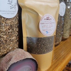 Puede incluir: Una bolsa de papel marr&oacute;n de t&eacute; Gentle Cortisol Support, con una ventana transparente que muestra las hojas de t&eacute;. La bolsa tiene una etiqueta circular con texto y est&aacute; junto a una piedra de &aacute;gata pulida y cortada con bandas rosas y blancas.
