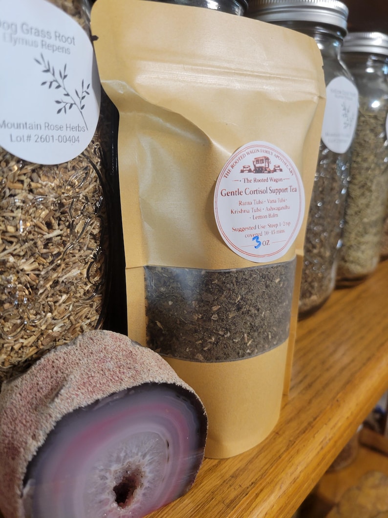 Puede incluir: Una bolsa de papel marr&oacute;n de t&eacute; Gentle Cortisol Support, con una ventana transparente que muestra las hojas de t&eacute;. La bolsa tiene una etiqueta circular con texto y est&aacute; junto a una piedra de &aacute;gata pulida y cortada con bandas rosas y blancas.