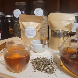 Puede incluir: Una exhibici&oacute;n de productos de t&eacute; de hierbas. La imagen presenta tazas de t&eacute; de vidrio llenas de t&eacute;, un peque&ntilde;o cuenco blanco y una pila de hojas de t&eacute; sueltas. Varios frascos y bolsas de papel de hierbas est&aacute;n en el fondo. El texto en la bolsa dice "The Rooted Wagon Gentle Cortisol Support Tea."