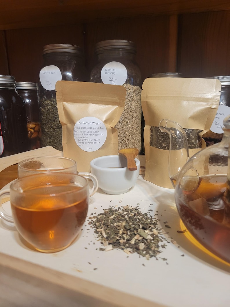 Puede incluir: Una exhibici&oacute;n de productos de t&eacute; de hierbas. La imagen presenta tazas de t&eacute; de vidrio llenas de t&eacute;, un peque&ntilde;o cuenco blanco y una pila de hojas de t&eacute; sueltas. Varios frascos y bolsas de papel de hierbas est&aacute;n en el fondo. El texto en la bolsa dice "The Rooted Wagon Gentle Cortisol Support Tea."