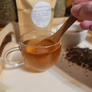 Puede incluir: Una taza de t&eacute; transparente llena de t&eacute; color &aacute;mbar, colocada sobre una bandeja blanca. Una cuchara de madera revuelve el t&eacute;. Una bolsa de papel marr&oacute;n con texto est&aacute; en el fondo, junto con hojas de t&eacute; sueltas. Escena de cata de t&eacute;.