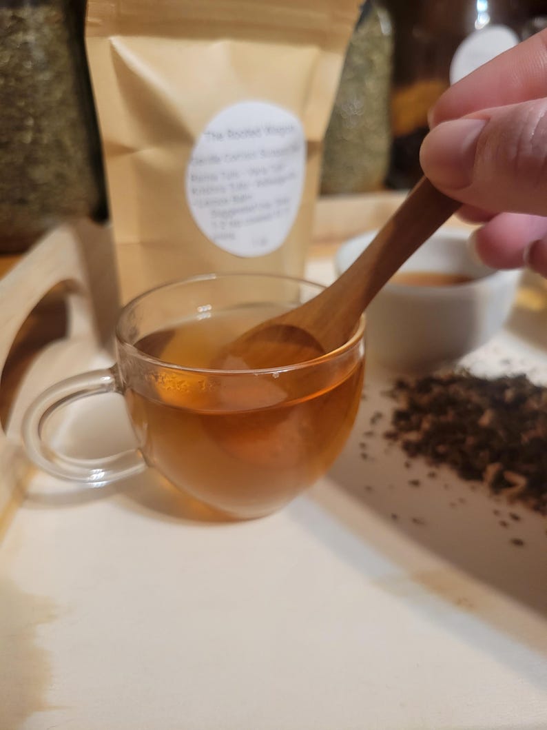Puede incluir: Una taza de t&eacute; transparente llena de t&eacute; color &aacute;mbar, colocada sobre una bandeja blanca. Una cuchara de madera revuelve el t&eacute;. Una bolsa de papel marr&oacute;n con texto est&aacute; en el fondo, junto con hojas de t&eacute; sueltas. Escena de cata de t&eacute;.