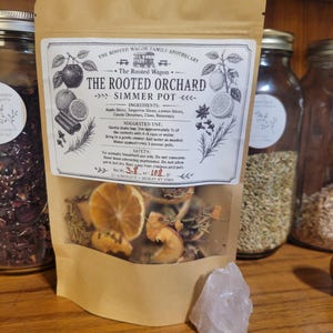 May include: A tan pouch labeled The Rooted Orchard Simmer Pot containing dried apple slices, tangerine slices, lemon slices, cinnamon sticks, cloves, and rosemary. The label lists ingredients and instructions. Net weight is 3.8 ounces or 108 grams.