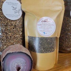 Puede incluir: Una bolsa de papel color beige con t&eacute; y una ventana transparente, etiquetada como "Gentle Cortisol Support Tea". Una geoda grande, pulida, rosa y blanca se encuentra frente a la bolsa. Un tarro de hierbas secas es visible en el fondo.