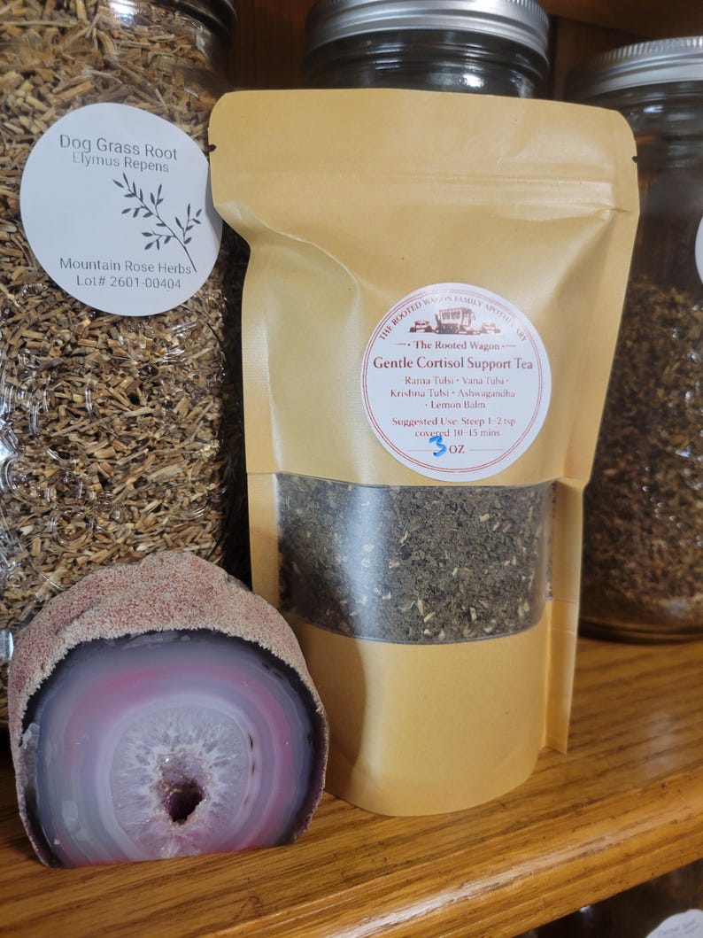 Puede incluir: Una bolsa de papel color beige con t&eacute; y una ventana transparente, etiquetada como "Gentle Cortisol Support Tea". Una geoda grande, pulida, rosa y blanca se encuentra frente a la bolsa. Un tarro de hierbas secas es visible en el fondo.