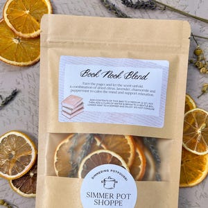 Peut inclure: Un sachet transparent de pot-pourri avec des tranches d'orange séchées, de la lavande et d'autres herbes. Le sachet porte les inscriptions "Book Nook Blend" et "Simmer Pot Shoppe". Des tranches d'orange séchées et des brins de lavande sont disposés autour du sachet.