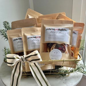 Peut inclure: Un panier doré rempli de plusieurs sachets en papier kraft étiquetés "Holiday Home". Chaque sachet contient des fruits secs et des épices. Un nœud rayé crème et vert orne le panier, décoré de brins de conifères.