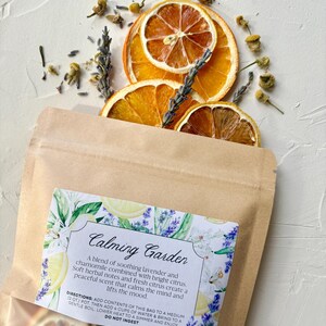 May include: A brown paper bag with a label reading "Calming Garden" is surrounded by dried orange slices, lavender sprigs, and chamomile flowers. The label describes a blend of lavender, chamomile, and citrus.