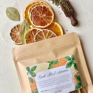 May include: A kraft paper bag labelled "Fresh Start Simmer" is surrounded by dried orange and lemon slices, bay leaves, and a wooden scoop of herbs. The label describes a refreshing blend of citrus, cool mint, and bay leaf.