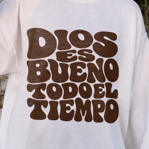 Puede incluir: Una sudadera blanca de manga larga con cuello redondo que presenta el texto en marrón oscuro DIOS ES BUENO TODO EL TIEMPO en la parte delantera con una tipografía gruesa, redondeada y de estilo retro. Tiene cuello, puños y dobladillo acanalados.