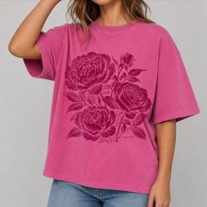May include: A fuchsia pink short-sleeve t-shirt featuring a dark magenta graphic print of blooming peonies and roses. The design includes the word still and the text Psalm 46:10 integrated within the floral illustration on the front.