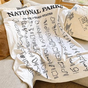 May include: A white throw blanket featuring a black printed map design of the National Parks of the United States. The fabric displays the text NATIONAL PARKS OF THE UNITED STATES at the top, followed by a grid of various park outlines and names.