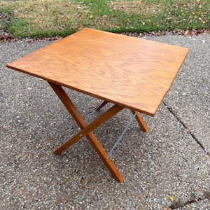 Puede incluir: Una mesa plegable cuadrada de madera con acabado marrón claro. La mesa tiene un diseño clásico con una estructura de patas en forma de X y una barra de soporte metálica. La mesa mide aproximadamente 61 cm de largo y de ancho.