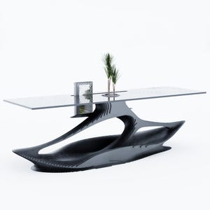 May include: A long rectangular table with a clear glass top and a dark, layered, organic-shaped base. The table features a central cutout containing a small green plant and a book with the word FOREST printed on the cover, resting on the glass.