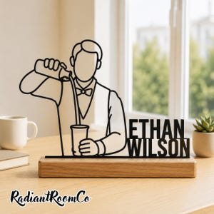 Peut inclure: Une sculpture en fil de métal noir représentant un barman versant une boisson dans un verre, montée sur une base rectangulaire en bois. La sculpture comporte le texte ETHAN WILSON en lettres capitales grasses intégré sur le côté droit.