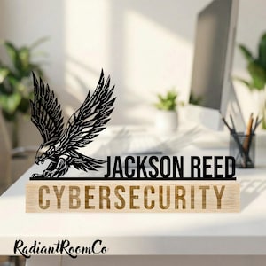 May include: A black metal eagle sculpture with outstretched wings, above the name "JACKSON REED" and the word "CYBERSECURITY" on a wooden base. The desk decor is set on a white surface.