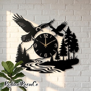 Peut inclure: Une horloge murale en métal noir représentant la silhouette de deux corbeaux en vol au-dessus d'un paysage forestier avec une rivière sinueuse. Le cadran circulaire possède des aiguilles dorées et des chiffres blancs aux positions 3, 6, 9 et 12.