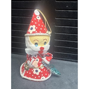Puede incluir: Adorno navideño vintage de Papá Noel. El adorno presenta un gorro y un vestido de lunares rojos y blancos con detalles de flores blancas. El Papá Noel tiene una cara amarilla, barba blanca y una cuerda dorada para colgar. Un pequeño árbol verde se sostiene en una mano.