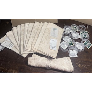 May include: A set of cream-coloured damask napkins and clear, faceted napkin rings. The napkins are stacked and folded, with a floral pattern. The napkin rings are round and have a textured design. Some napkins have price tags.