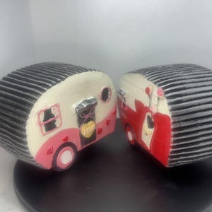 May include: Two decorative camper figurines. One is cream and pink with a black roof, the other is red and cream with a black roof. Both have heart accents and a door with a heart-shaped "Love" sign.