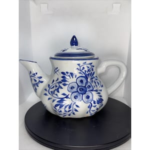 May include: White ceramic teapot with a blue floral design. The teapot features a curved spout, a rounded body, and a curved handle. The lid is topped with a small blue finial. The teapot is decorated with blue floral and leaf patterns.