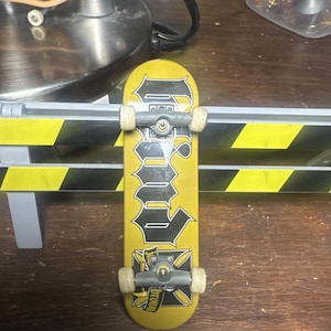 Pode incluir: Um fingerboard amarelo e preto com a palavra "Ground" em letras grandes e estilizadas. O skate em miniatura tem eixos prateados e rodas brancas. O deck apresenta um desenho de cruz na parte inferior.