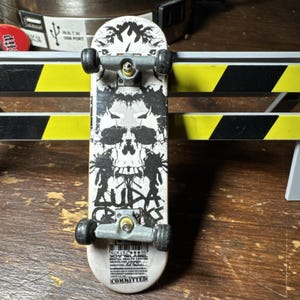 Puede incluir: Una pequeña fingerboard blanca con un gráfico de calavera negra y el texto "AUDA CROOKS". El monopatín en miniatura tiene ruedas negras y ejes plateados. La parte inferior de la tabla tiene el texto "COMMITTED".