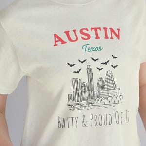 May include: Cream-colored t-shirt with the text "AUSTIN Texas" in red and teal above a black and white skyline graphic. Below the graphic, the text reads "BATTY & PROUD OF IT".