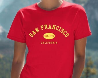 May include: A red t-shirt featuring the text "SAN FRANCISCO" in yellow, arched above an oval with "EST. 1776" and "CALIFORNIA" below. The shirt is worn by a person against a mountain backdrop.