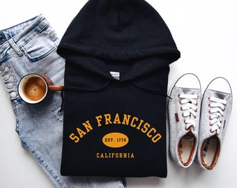 Op de afbeelding: Zwarte hoodie met de tekst "SAN FRANCISCO EST. 1776 CALIFORNIA" in goudkleurige letters. De afbeelding toont ook een lichtblauwe jeans, grijze sneakers en een koffiemok.
