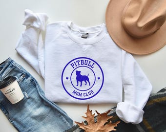 Puede incluir: Una sudadera blanca con un diseño circular azul que dice "PITBULL MOM CLUB" con la silueta de un perro. La imagen incluye vaqueros azules, una taza de café y un sombrero color canela.