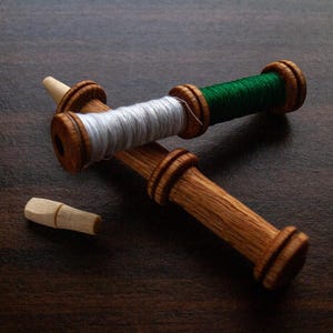 Puede incluir: Tres carretes de hilo de madera con hilo blanco y verde, y una pequeña clavija de madera. Los carretes son de madera marrón claro y están dispuestos sobre una superficie marrón oscuro. Los carretes de hilo se utilizan para coser y hacer manualidades.
