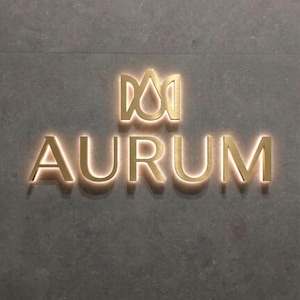 May include: Gold-colored metal sign with the word "AURUM" in capital letters, mounted on a gray textured wall. The sign has a stylized crown or emblem above the word, and the letters are illuminated with a warm glow.
