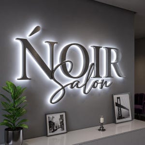 May include: A wall-mounted 3D sign featuring the text 'NOIR Salon' in dark metallic lettering. The sign is backlit with a bright white glow that creates a halo effect against the gray wall. The letters have a clean, professional finish.