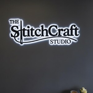 May include: The StitchCraft Studio logo, a black and white sign, is mounted on a gray wall. The text is in a stylized font with a white neon glow around the letters. The word "The" is above "StitchCraft", and "STUDIO" is below.
