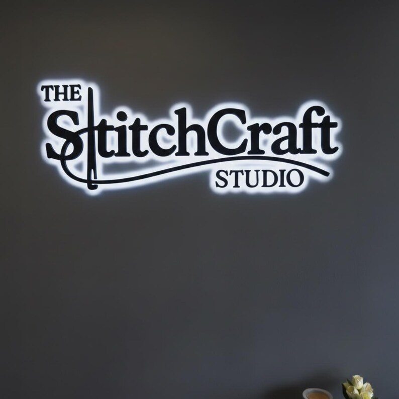 May include: The StitchCraft Studio logo, a black and white sign, is mounted on a gray wall. The text is in a stylized font with a white neon glow around the letters. The word "The" is above "StitchCraft", and "STUDIO" is below.