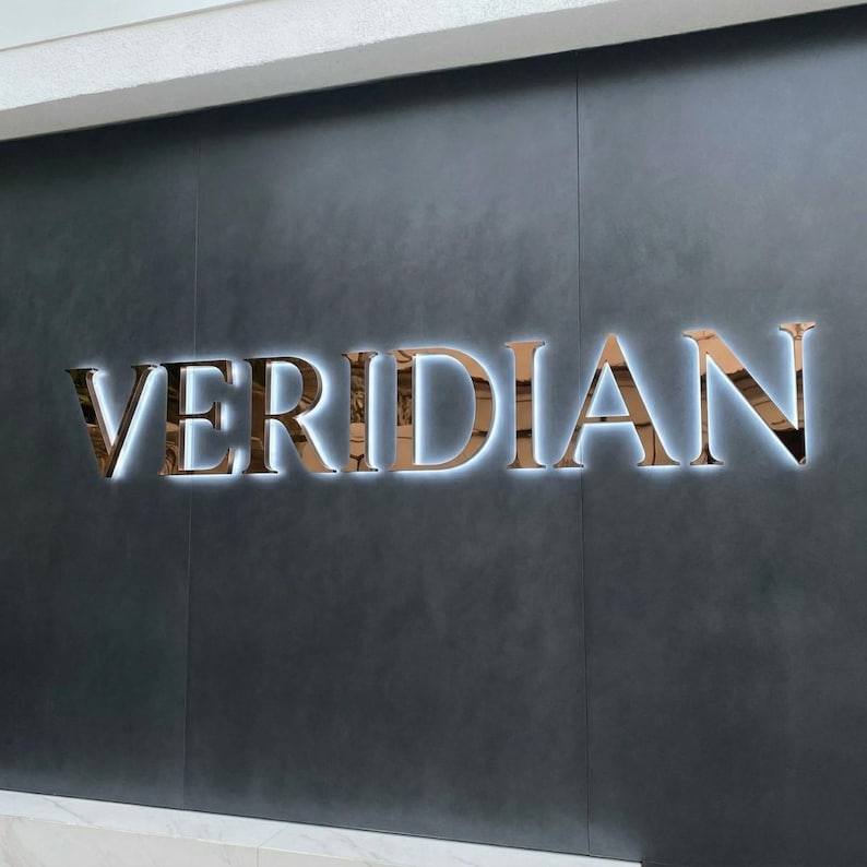 May include: The word "VERIDIAN" in large, metallic, copper-colored letters is mounted on a dark gray wall. The letters are backlit with a bright white light, creating a striking contrast. The sign is likely for a business or brand.