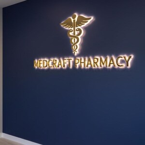 May include: A navy blue wall features a gold-colored pharmacy logo and the words "MEDCRAFT PHARMACY" in gold lettering. The logo is a caduceus symbol with a pair of wings above a staff and two snakes. The sign is illuminated.