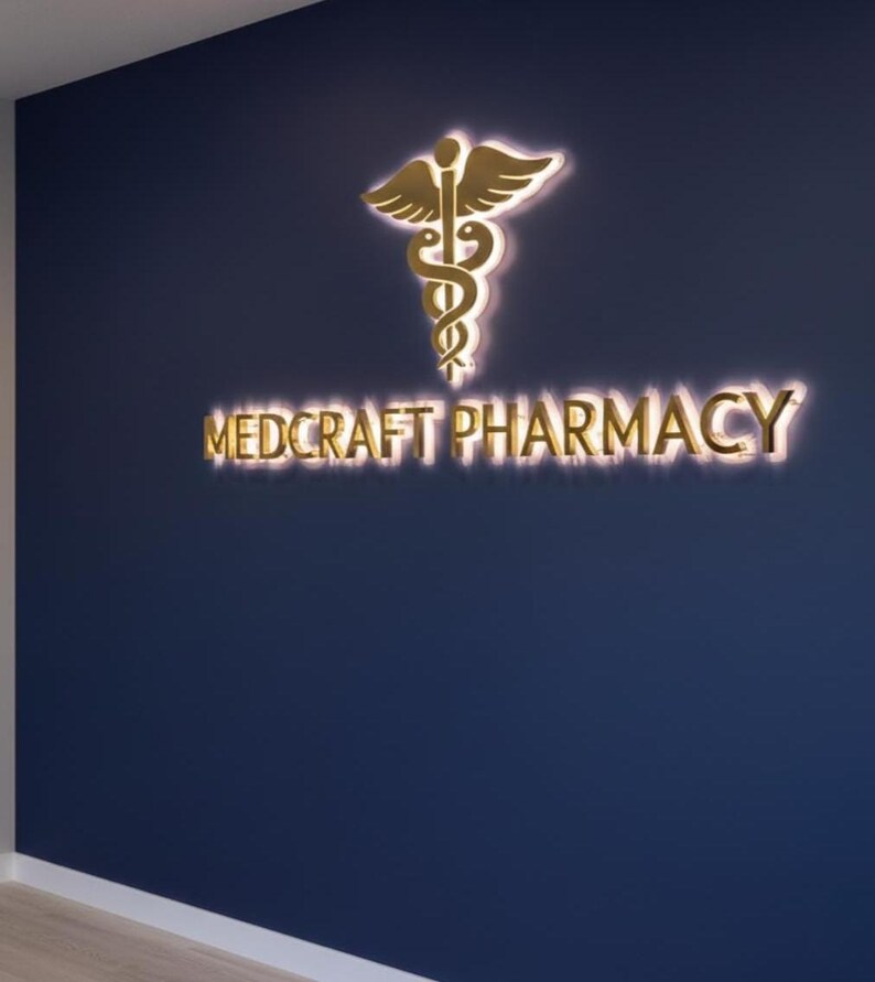 May include: A navy blue wall features a gold-colored pharmacy logo and the words "MEDCRAFT PHARMACY" in gold lettering. The logo is a caduceus symbol with a pair of wings above a staff and two snakes. The sign is illuminated.