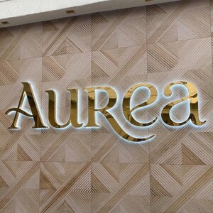 May include: Gold-colored "Aurea" sign with a white backlight, mounted on a textured, light brown wall. The sign's elegant, stylized font is illuminated, creating a striking visual effect. The wall has a geometric pattern.