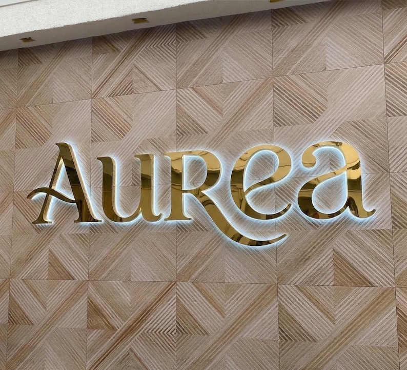 May include: Gold-colored "Aurea" sign with a white backlight, mounted on a textured, light brown wall. The sign's elegant, stylized font is illuminated, creating a striking visual effect. The wall has a geometric pattern.