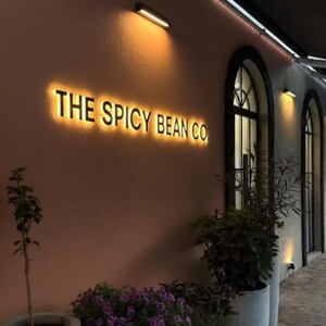 May include: Exterior of a cafe with the illuminated text "THE SPICY BEAN CO." on a salmon-colored wall. The scene includes arched windows, potted plants with purple flowers, and warm lighting, creating an inviting atmosphere.