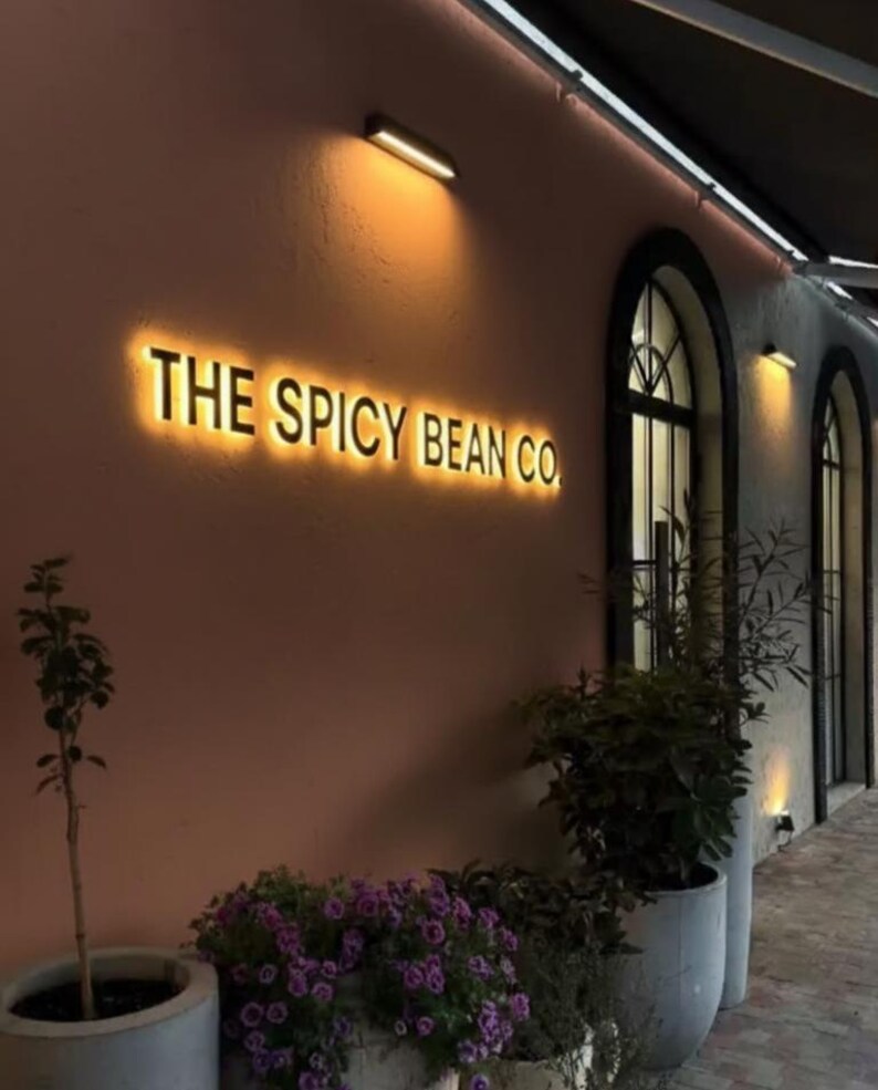 May include: Exterior of a cafe with the illuminated text "THE SPICY BEAN CO." on a salmon-colored wall. The scene includes arched windows, potted plants with purple flowers, and warm lighting, creating an inviting atmosphere.