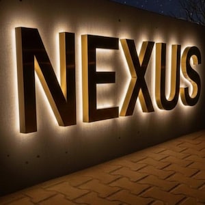 以下が含まれることがあります： 大きな金色のブロック文字で「NEXUS」という単語を表示する照明付きの看板。文字はコンクリートの壁に取り付けられ、各文字を縁取る暖かく光る光があります。背景は夜のシーンです。