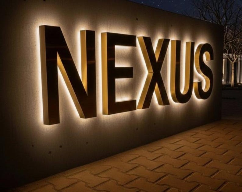 May include: Illuminated sign spelling "NEXUS" in large, gold-colored block letters. The letters are mounted on a dark gray wall with a warm, glowing light outlining each letter. The sign is outdoors, with a brick walkway in the foreground.