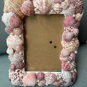 May include: A rectangular white picture frame decorated with various seashells in shades of pink, cream, and white. Small faux pearls are interspersed among the shells. The frame has a brown backing.