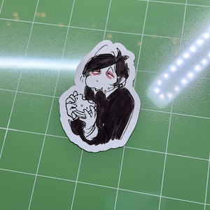 Puede incluir: Una pegatina con una ilustración en blanco y negro de una persona sosteniendo un sándwich. La persona tiene el pelo negro y viste un atuendo negro. La pegatina tiene un borde blanco.