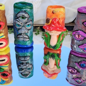 May include: Four colorful, hand-painted ceramic tiki-style sculptures. The sculptures feature vibrant colors and unique designs, including faces, mushrooms, and eyes. The sculptures are displayed on a reflective surface.