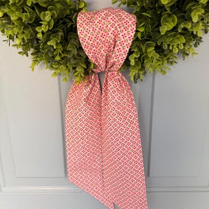 May include: A green faux-leaf wreath with a pink floral patterned bow. The bow is made of fabric with a repeating pattern of small red flowers and green leaves on a white background. The wreath is hanging on a light blue door.