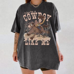 May include: A charcoal grey short-sleeve t-shirt featuring a graphic of a cartoon cowboy riding a horse. The text "You're a COWBOY LIKE ME" is printed in a light beige font above and below the illustration, which includes cacti and desert scenery.